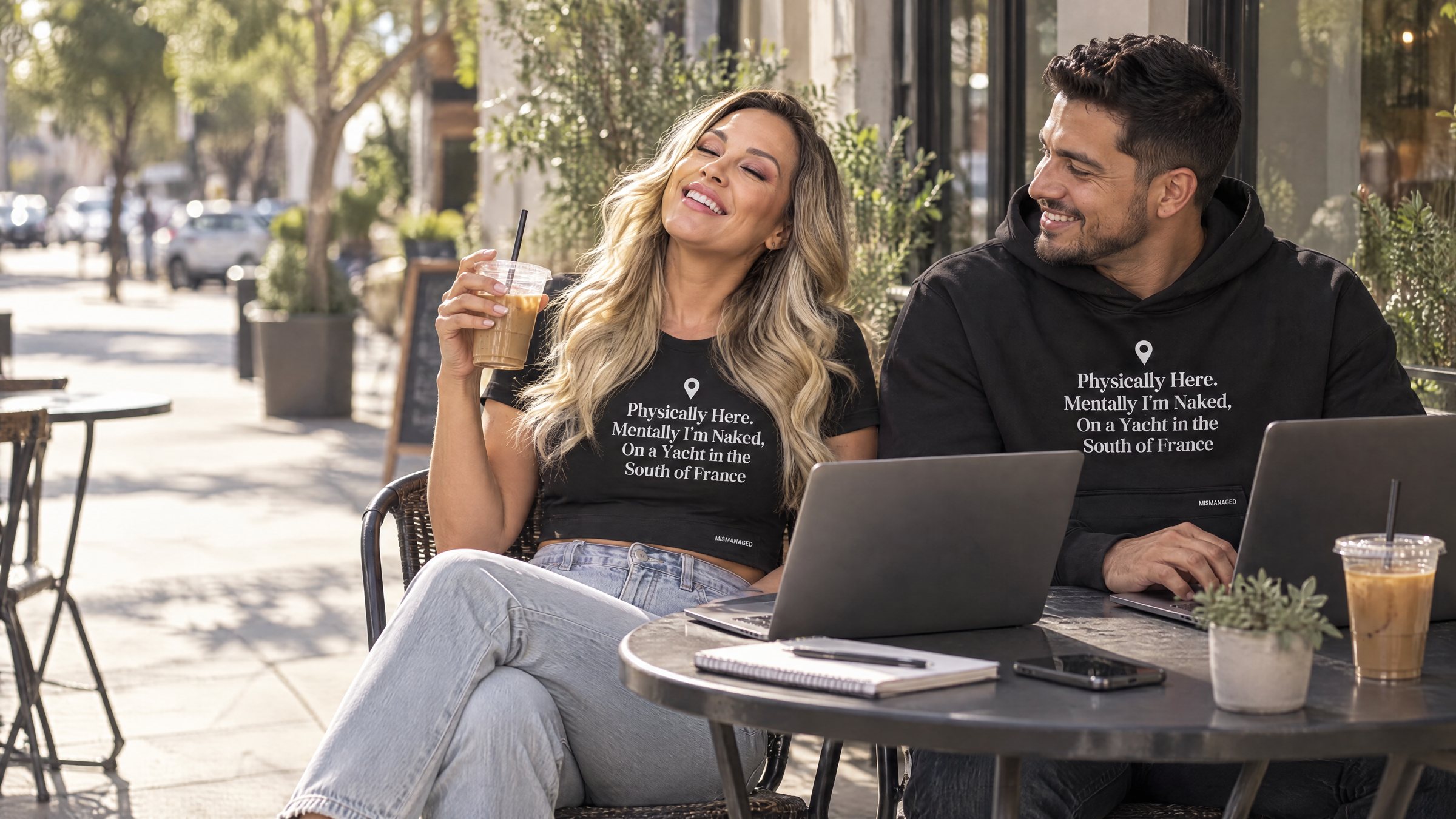 Two people working at a sunny cafe terrace wearing the South of France crop tee and hoodie